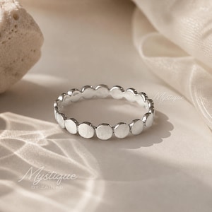 May include: A silver ring with a repeating dot design. The ring is circular and features a series of small, evenly spaced, flat circles around its circumference. The ring is displayed on a neutral-toned surface.