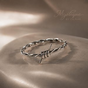 May include: A silver-toned ring featuring a twisted band design that mimics the appearance of barbed wire, with a central coiled section. The text Mystique BY ZAINAB is visible in the upper right corner of the image, with a smaller watermark.