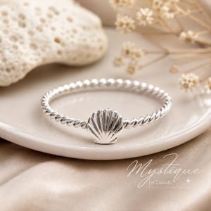 May include: A silver ring featuring a twisted band design with a central seashell charm. The metal has a polished finish, and the ring is positioned on a smooth surface. The text Mystique by Zainab is visible in the lower right corner of the image.