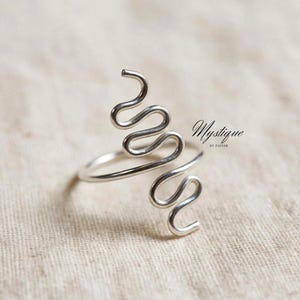 May include: A silver-toned metal ring featuring a continuous wire design that forms two vertical, serpentine, or zigzag patterns extending upwards and downwards from the central band. The text Mystique BY ZAINAB is printed in black to the right.