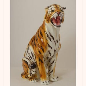 May include: A ceramic tiger statue with an open mouth, showcasing its teeth and pink interior. The tiger has orange and black stripes, white underbelly, and is posed in a seated position. The statue is a decorative item.