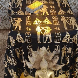 Puede incluir: Un mantel negro con jeroglíficos y figuras egipcias doradas, con una estatua de Buda dorada y una baraja de cartas del tarot. El mantel está drapeado sobre una mesa, creando una atmósfera mística.
