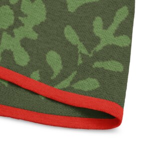 May include: Close-up of a green camouflage pattern fabric with a red border. The fabric has a textured appearance and features a repeating pattern of green leaf-like shapes on a darker green background. The red border adds a contrasting accent.