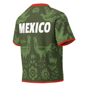 May include: Olive green t-shirt with red trim and the word "MEXICO" in white block letters. The shirt features a repeating pattern of green birds, flowers, and other decorative elements.