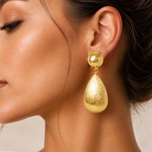 May include: A gold-toned drop earring featuring a textured spherical stud connected to a larger, elongated teardrop-shaped pendant. The surface of the metal has a brushed, matte finish with visible fine lines, creating a reflective, metallic appearance.