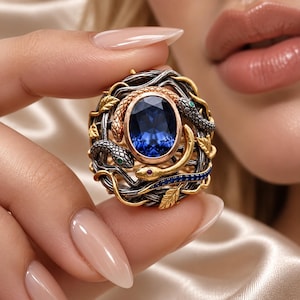 May include: A ring featuring a large oval-cut deep blue gemstone set in a rose gold bezel. The band is crafted from oxidized silver and gold-toned metal, intricately shaped like intertwined branches, leaves, and three small snakes with tiny colored eyes.