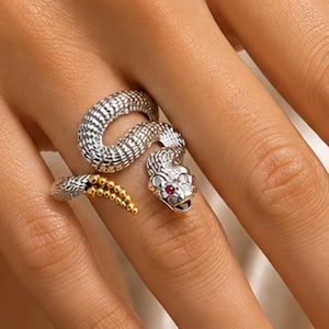 May include: A silver-toned snake ring featuring a textured, scaled body that wraps around the finger. The snake head is detailed with a small, vibrant red gemstone eye, while the tail end is accented with a series of polished gold-toned beads.