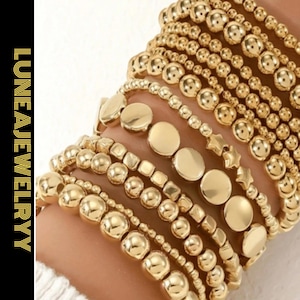 May include: A collection of gold beaded bracelets in various styles, including round beads, flat disc beads, and star-shaped beads. The bracelets are stacked on a wrist, showcasing the different designs. The brand name "LUNEAJEWELRY" is displayed vertically on the left.