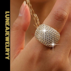 Könnte beinhalten: Ein goldfarbener Ring mit einer großen, abgerundeten Oberfläche, dicht bedeckt mit kleinen, funkelnden, klaren Steinen. Der Ring befindet sich an einem Finger. Der Text "LUNEA JEWELRY" ist vertikal auf schwarzem Hintergrund ausgerichtet.