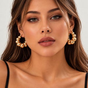 May include: A pair of gold hoop earrings featuring a graduated design with multiple polished spherical beads of varying sizes arranged along the circular wire frame. The earrings have a high-shine metallic finish and a secure clasp mechanism.
