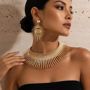 May include: A gold-toned metal choker necklace featuring a series of radiating, thin, pointed spikes arranged in a circular collar design, paired with matching circular sunburst-style drop earrings with a central disc and thin rays.
