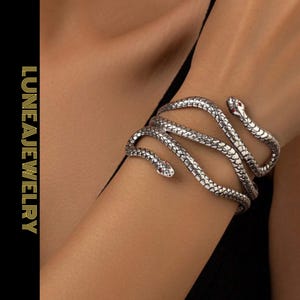 May include: A silver-toned snake cuff bracelet with red gemstone eyes wraps around a wrist. The bracelet has a textured, scale-like pattern. The jewelry is from LUNEAJEWELRY.