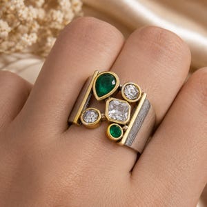 May include: A wide band ring featuring a textured silver finish and gold-tone accents. The centre is adorned with a cluster of five stones: two pear-shaped and round green gems, and three clear, faceted white stones in square and round shapes.