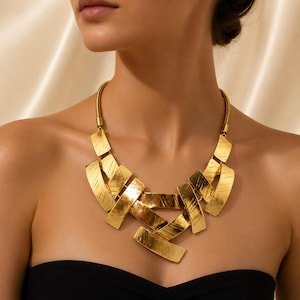 May include: A gold-toned statement necklace featuring a series of interconnected, rectangular metal plates with a brushed, textured finish. The plates are arranged in an overlapping, geometric pattern, suspended from a flexible, rounded snake chain.