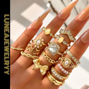Könnte beinhalten: Eine Hand zeigt eine Sammlung goldener Ringe, darunter ein Ring in Schleifenform, ein Ring mit einem großen, klaren Stein und Ringe mit Perlen. Die Nägel sind lang und mit einem Nude- und Weiß-Design bemalt. Der Text "LUNEAJEWELRYY" befindet sich links.