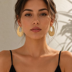 May include: A pair of gold-toned hoop earrings featuring a distinct sunburst or fan-like pleated texture. The earrings have an elongated oval shape with a polished metallic finish, catching the light across the radiating ridges of the metal surface.