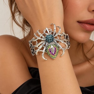 Op de afbeelding: Een zilverkleurige armband met een spinnenontwerp. Het lichaam van de spin is ingelegd met kleine blauwgroene, paarse, limoengroene en gele strass-steentjes, met een heldere marquise-geslepen steen in het midden van het achterlijf.