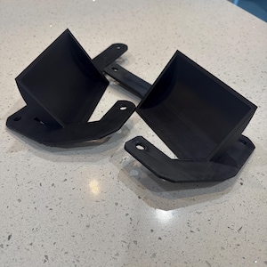 Puede incluir: Dos soportes negros impresos en 3D con una copa cuadrada y una base plana con un agujero. Los soportes están diseñados para sujetar un objeto. Los artículos están sobre una encimera moteada.