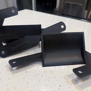 May include: Two black metal brackets with rounded edges and mounting holes. One bracket is folded, and the other is a rectangular shape. The text "CarsNKirby" is visible on one of the brackets.
