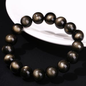 May include: A black and gold beaded bracelet. The bracelet is made up of numerous round beads with a glossy finish and a speckled, golden shimmer. The beads are strung together, forming a circular shape, resting on a white surface.