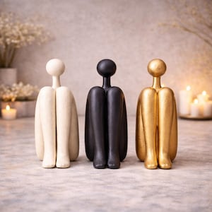 May include: Three abstract human figure sculptures in cream, black, and gold. Each figure is seated with legs drawn up, and a rounded head. The sculptures are displayed on a light-colored surface, with candles and flowers in the background.