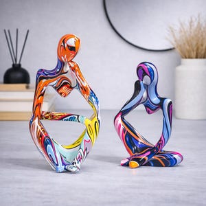 Op de afbeelding: Twee abstracte sculpturen in zittende houding. De sculptuur aan de linkerkant is beschilderd met oranje, gele, blauwe en witte wervelingen. De sculptuur aan de rechterkant is beschilderd met blauwe, roze en paarse wervelingen. Beide zijn decoratieve kunstwerken.