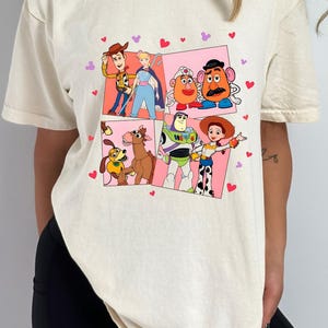 May include: Cream-colored t-shirt featuring a colorful graphic of characters from the animated film "Toy Story." The design includes Woody, Buzz Lightyear, Jessie, Mr. Potato Head, and others, set against pink and white panels with heart accents.