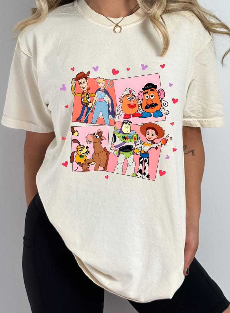 May include: Cream-colored t-shirt featuring a colorful graphic of characters from the animated film "Toy Story." The design includes Woody, Buzz Lightyear, Jessie, Mr. Potato Head, and others, set against pink and white panels with heart accents.