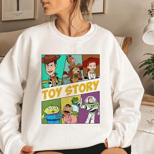 May include: White sweatshirt featuring a colorful graphic of Toy Story characters. The design includes Woody, Buzz Lightyear, and other characters from the animated film, with the words "TOY STORY" in yellow.