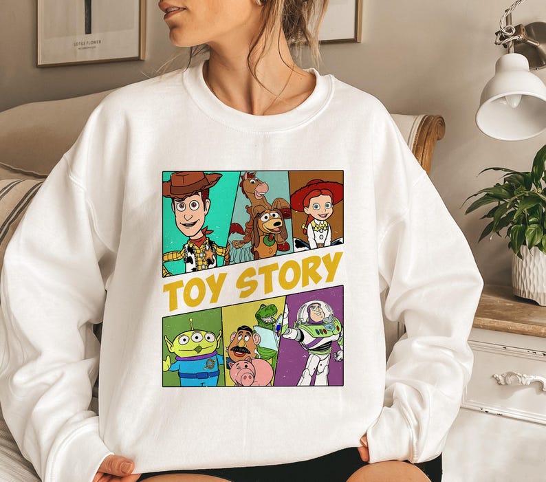 May include: White sweatshirt featuring a colorful graphic of Toy Story characters. The design includes Woody, Buzz Lightyear, and other characters from the animated film, with the words "TOY STORY" in yellow.