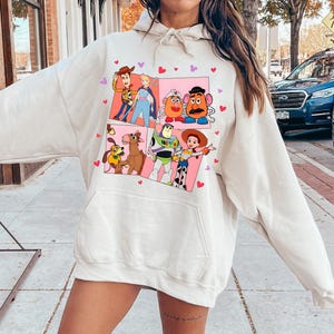May include: Cream-colored hoodie featuring a colorful graphic of characters from the animated movie Toy Story. The graphic is divided into panels with pink backgrounds and heart accents. The hoodie has a drawstring hood and a front pocket.