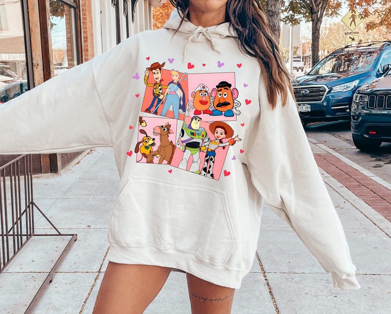 May include: Cream-colored hoodie featuring a colorful graphic of characters from the animated movie Toy Story. The graphic is divided into panels with pink backgrounds and heart accents. The hoodie has a drawstring hood and a front pocket.