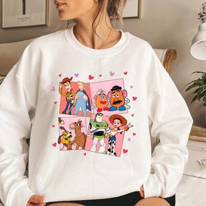 May include: White sweatshirt featuring a colorful graphic of Toy Story characters in a grid pattern. The design includes Woody, Buzz Lightyear, Jessie, and Mr. Potato Head, with pink and red heart accents. A cozy, casual top.