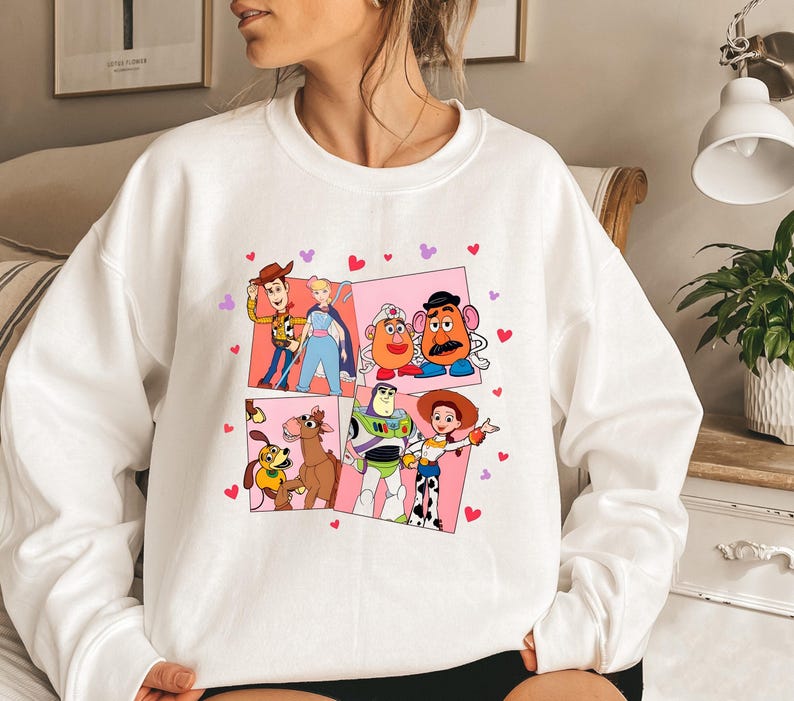 May include: White sweatshirt featuring a colorful graphic of Toy Story characters in a grid pattern. The design includes Woody, Buzz Lightyear, Jessie, and Mr. Potato Head, with pink and red heart accents. A cozy, casual top.