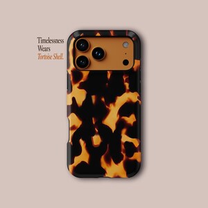 Op de afbeelding: Een telefoonhoesje met een schildpadpatroon in zwart en amber tinten. De hoes heeft een zwarte rand en een bruin camera gedeelte. De tekst "Timelessness Wears Tortoise Shell" staat links afgedrukt.