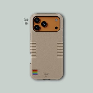 Puede incluir: Una funda de smartphone beige con superficie texturizada, que presenta un módulo de cámara ocre, agarres laterales acanalados y un pequeño icono cuadrado de arcoíris. El texto "Classic 1984" está impreso en negro abajo y a la izquierda.