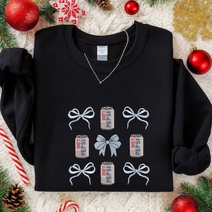 Puede incluir: Sudadera negra con latas de Diet Pepsi bordadas y lazos blancos. El diseño incluye tres latas de Diet Pepsi y seis lazos blancos, con un lazo plateado grande en el centro. Se ve un collar de oro.