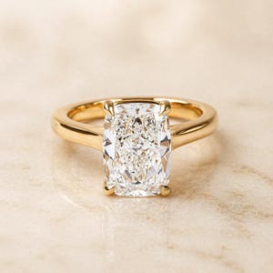 May include: A gold engagement ring featuring a rectangular-cut diamond. The diamond has rounded corners and is held in place by four prongs. The gold band is smooth and polished. The ring is presented on a light-toned surface, showcasing its brilliance.