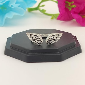 May include: A silver-colored ring with an open design featuring two detailed wing shapes. The ring is displayed on a black, hexagonal stand, with blurred flowers in the background.