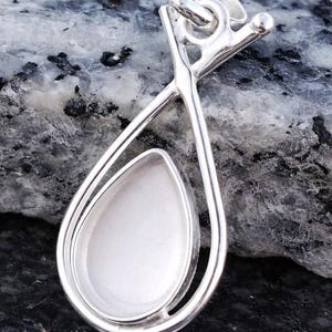 May include: A silver pendant with a teardrop-shaped setting. The pendant has a polished finish and a loop for a chain. The setting is designed to hold a stone or other decorative element. The pendant is displayed on a dark, speckled surface.