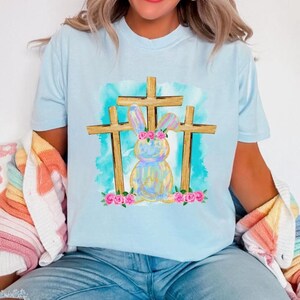 Könnte beinhalten: Hellblaues T-Shirt mit einem Aquarelldesign eines Hasen mit Blumenkrone, platziert zwischen drei Holzkreuzen. Der Hase ist in Pastellfarben gehalten, mit rosa Rosen an der Basis. Der Hintergrund hat eine blaue Aquarellwaschung.