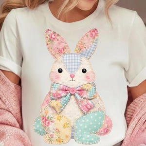 May include: White t-shirt with a patchwork bunny design. The bunny features pink, blue, and yellow floral and polka dot patterns, with a blue gingham centre. A pink bow with a blue centre adorns the bunny's neck, creating a whimsical and festive look.