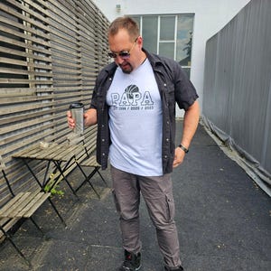 Op de afbeelding: Een man met een zonnebril, een zwart overhemd over een wit t-shirt met een basketbalafbeelding en het woord "PAPA" erop gedrukt. Hij draagt ​​grijze cargobroeken en zwarte sneakers. Hij houdt een zilveren reismok vast.