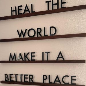 May include: A wall-mounted sign with the message "HEAL THE WORLD MAKE IT A BETTER PLACE." The black letters are mounted on dark brown wooden shelves against a white wall. The sign is designed to inspire and motivate.