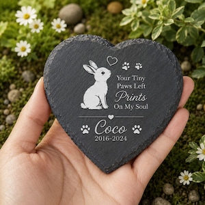 Może przedstawiać: Kamień pamiątkowy w kształcie serca z ciemnoszarego łupku. Po lewej stronie znajduje się ilustracja białego kr&oacute;lika z napisem "Your Tiny Paws Left Prints On My Soul" i imieniem "Coco" z datami 2016-2024. Kamień jest trzymany w dłoni.