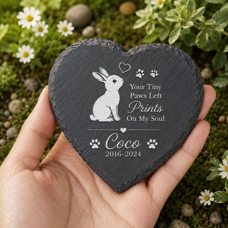 Może przedstawiać: Kamień pamiątkowy w kształcie serca z ciemnoszarego łupku. Po lewej stronie znajduje się ilustracja białego kr&oacute;lika z napisem "Your Tiny Paws Left Prints On My Soul" i imieniem "Coco" z datami 2016-2024. Kamień jest trzymany w dłoni.