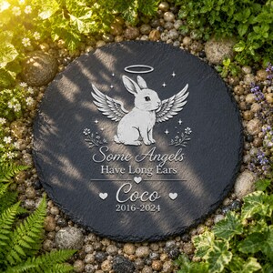 Może przedstawiać: Okrągły, ciemnoszary kamień pamiątkowy z białym wzorem anioła-kr&oacute;lika. Tekst brzmi "Some Angels Have Long Ears" i "Coco 2016-2024". Kamień otoczony jest zielenią i małymi kamieniami.