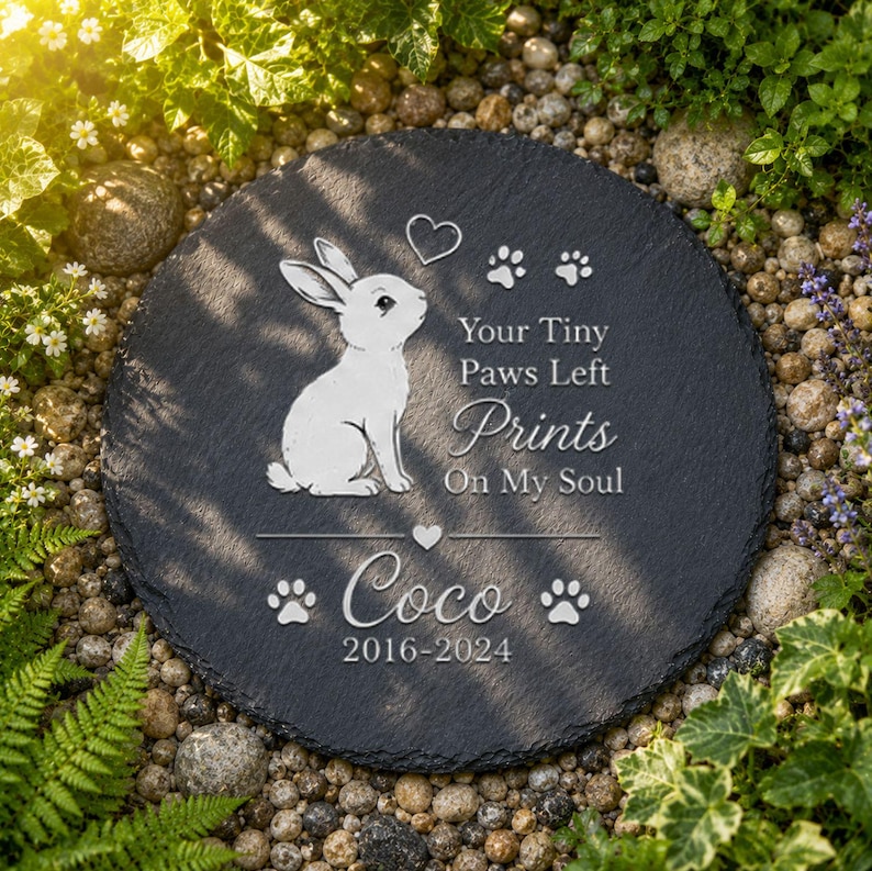 Może przedstawiać: Okrągły, ciemnoszary kamień pamiątkowy z białą ilustracją kr&oacute;lika i napisem "Your Tiny Paws Left Prints On My Soul". Imię "Coco" i lata "2016-2024" są r&oacute;wnież wyryte. Kamień otoczony jest małymi kamieniami i zielenią.