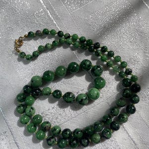 May include: A green beaded necklace with a gold clasp. The necklace features round beads in varying sizes, with dark green and black speckles. The necklace is displayed on a white, textured surface.