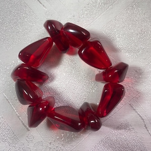 May include: A red beaded bracelet made of irregularly shaped, translucent beads. The beads are a deep red colour and appear to be strung together to form a circular shape. The bracelet is displayed on a white, textured surface.